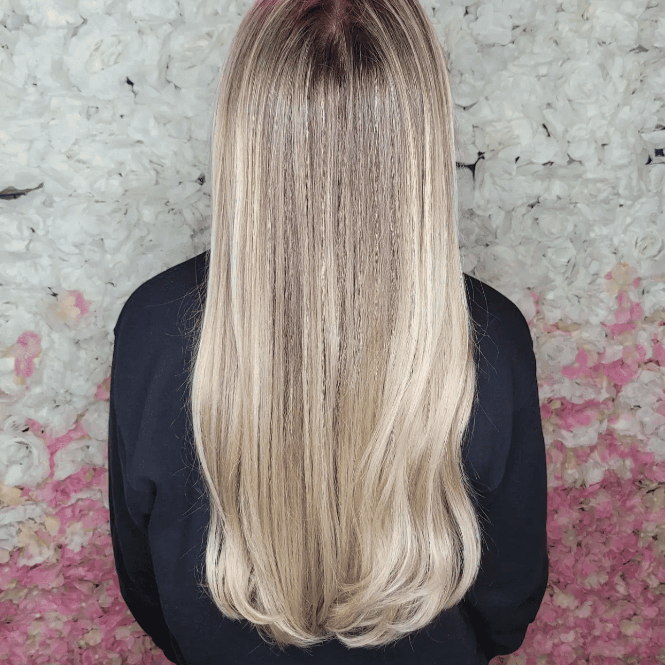 Person with long, straight blonde hair standing against a floral background.