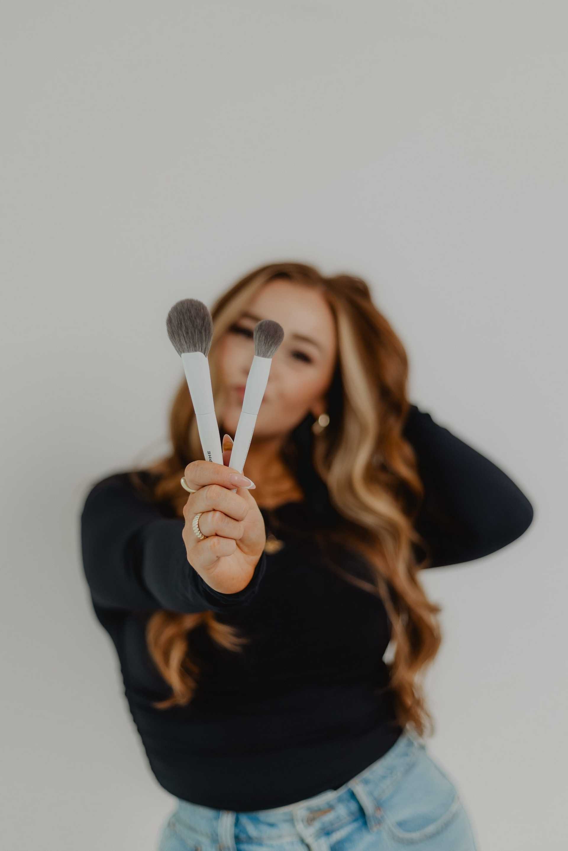 Woman holding makeup brushes, smiling, long wavy hair, casual outfit.